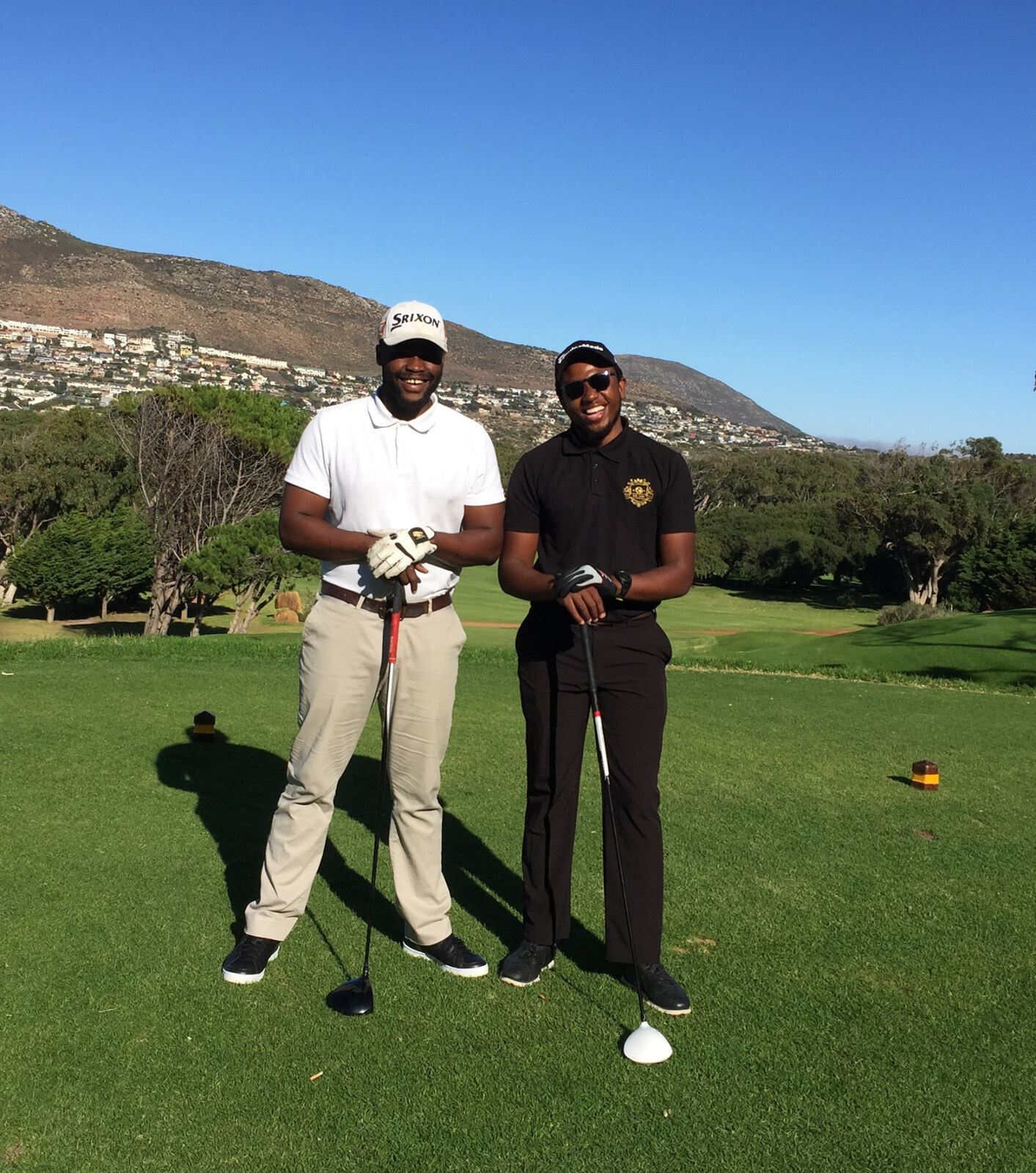 Tshepo and Mangezi on a tee box holding drivers, a mountain rising behind them
